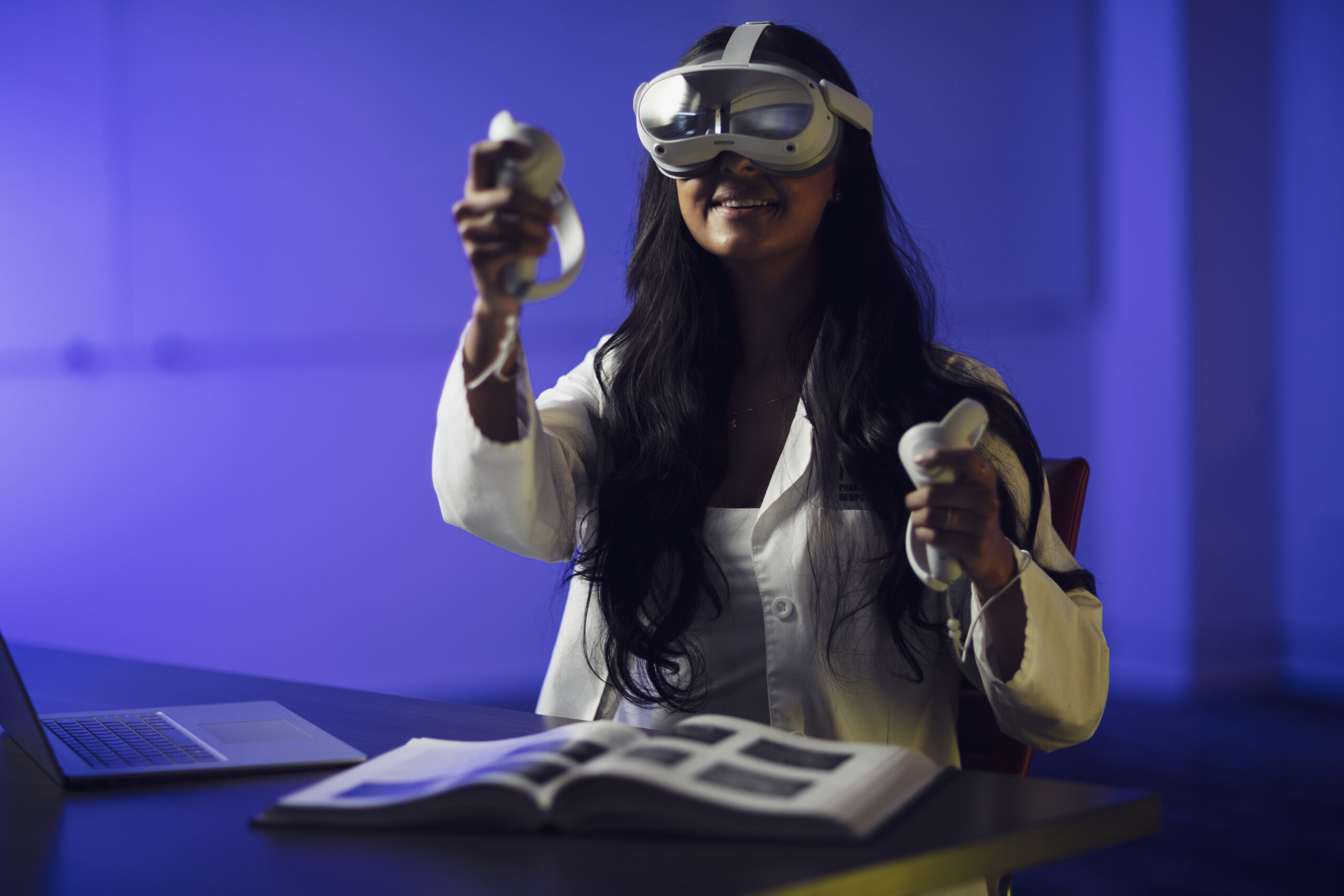 Woman in doctor's coat wearing a virtual reality headset with a radiology textbook in front of her.