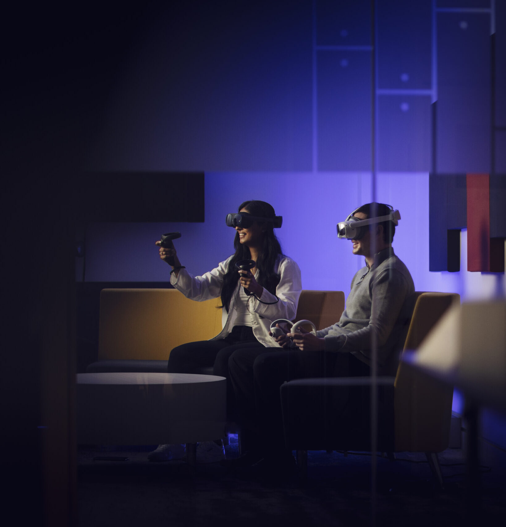 Woman in doctor's coat and man sitting on a couch together, both wearing virtual reality headsets.