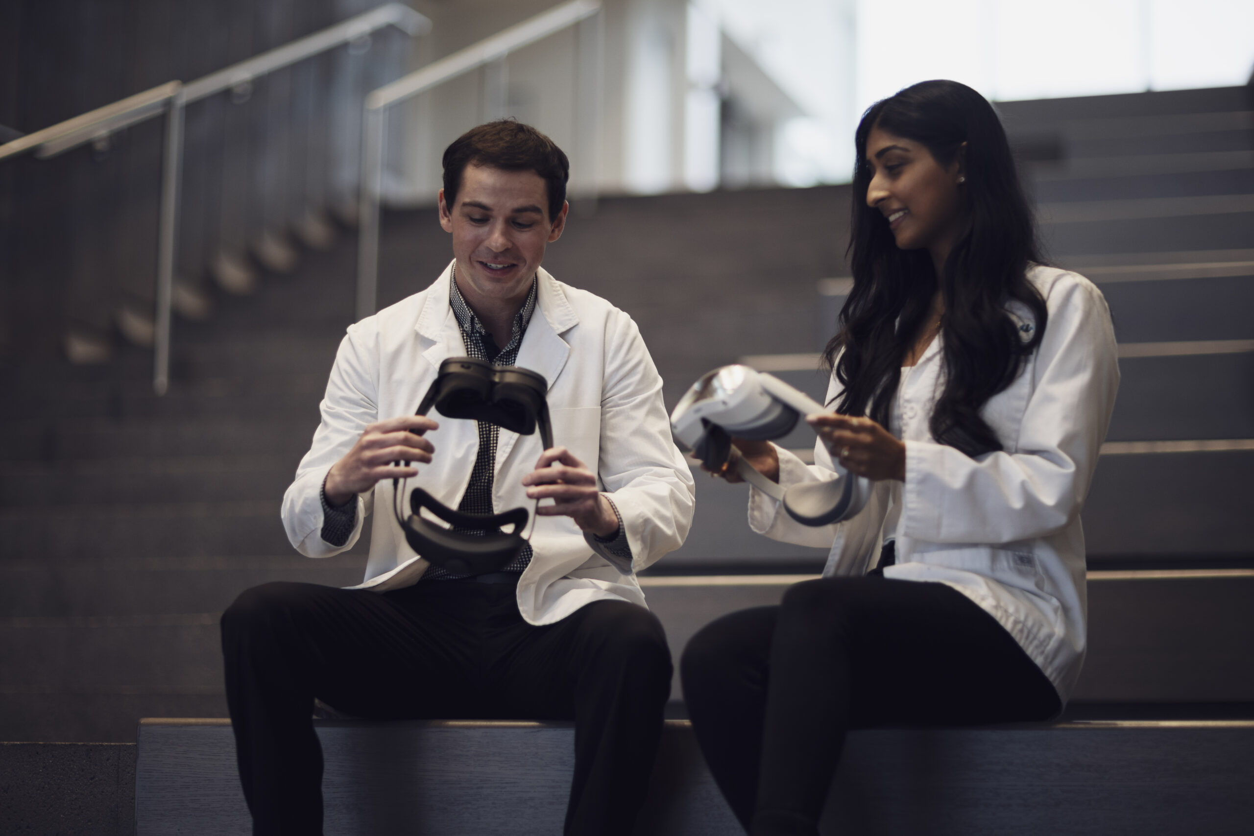 Woman in doctor's coat and man in doctor's coat sitting on a staircase together, both holding virtual reality headsets.