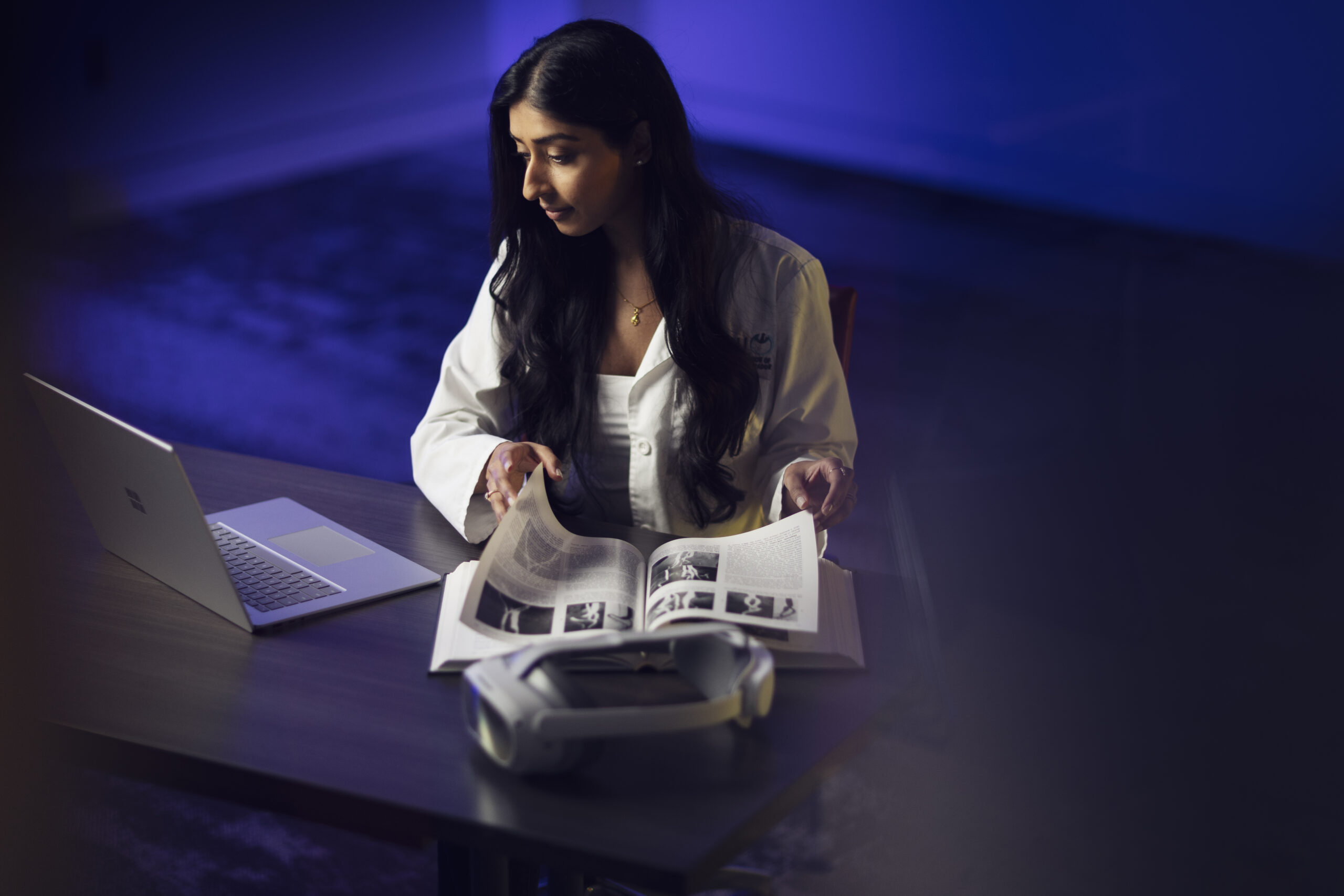 Woman in doctor's coat reading a medical imaging textbook with a virtual reality headset in front of her.