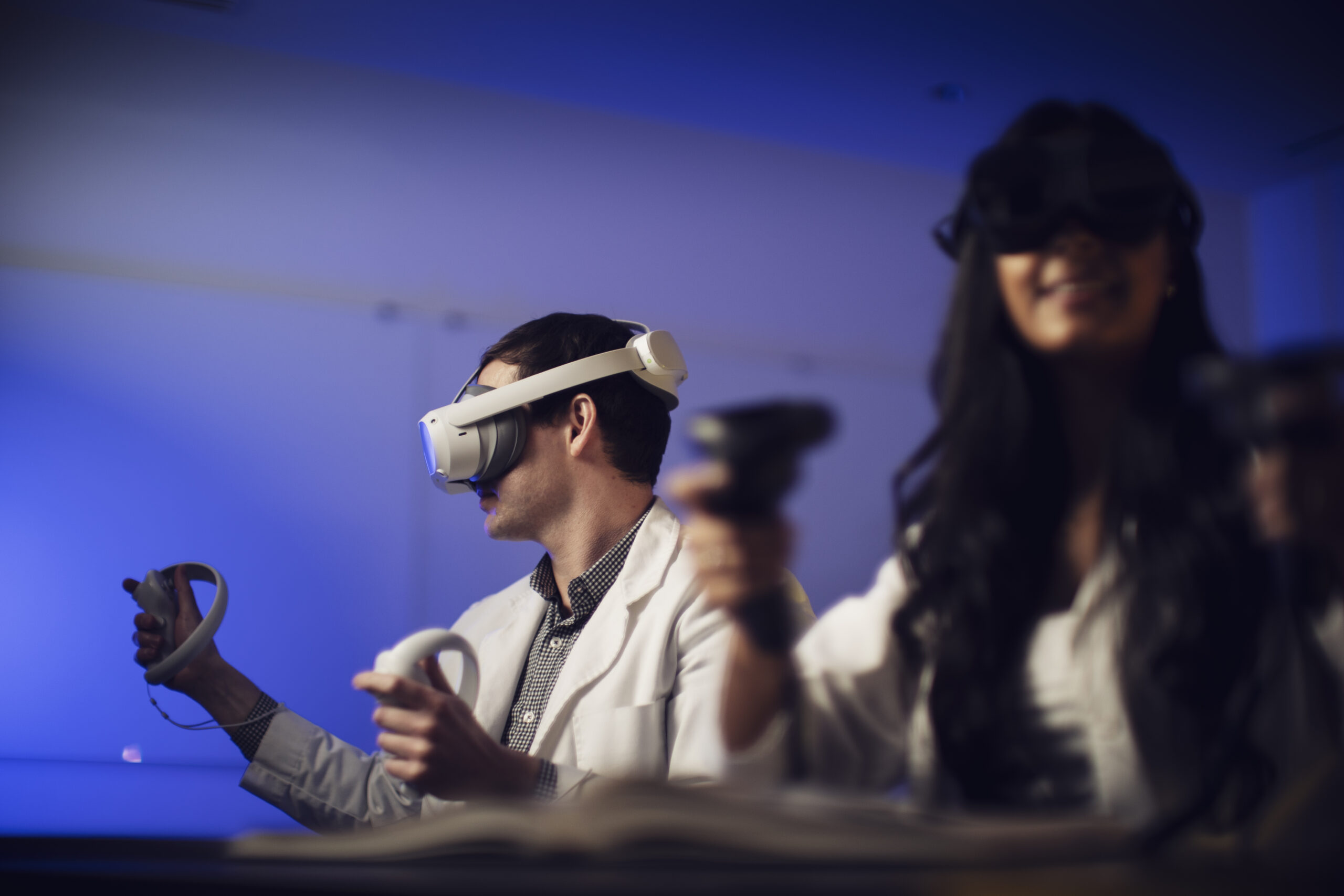 Woman in doctor's coat and man in doctor's sitting at a table together, both wearing virtual reality headsets.