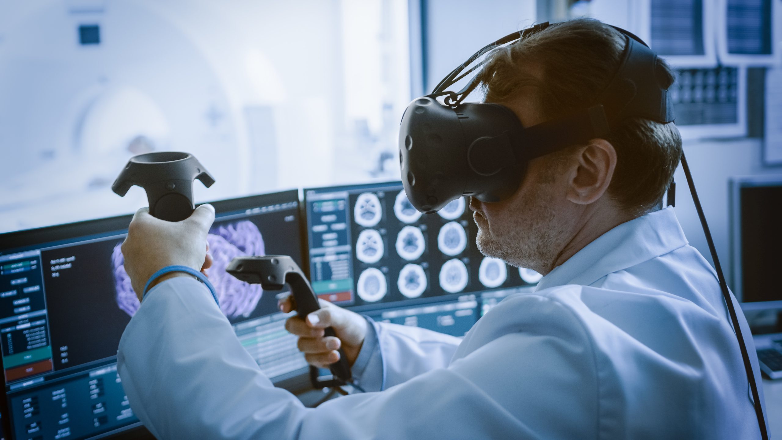 Radiologist wearing virtual reality headset, using controllers, and sitting in front of 2D radiology set-up.
