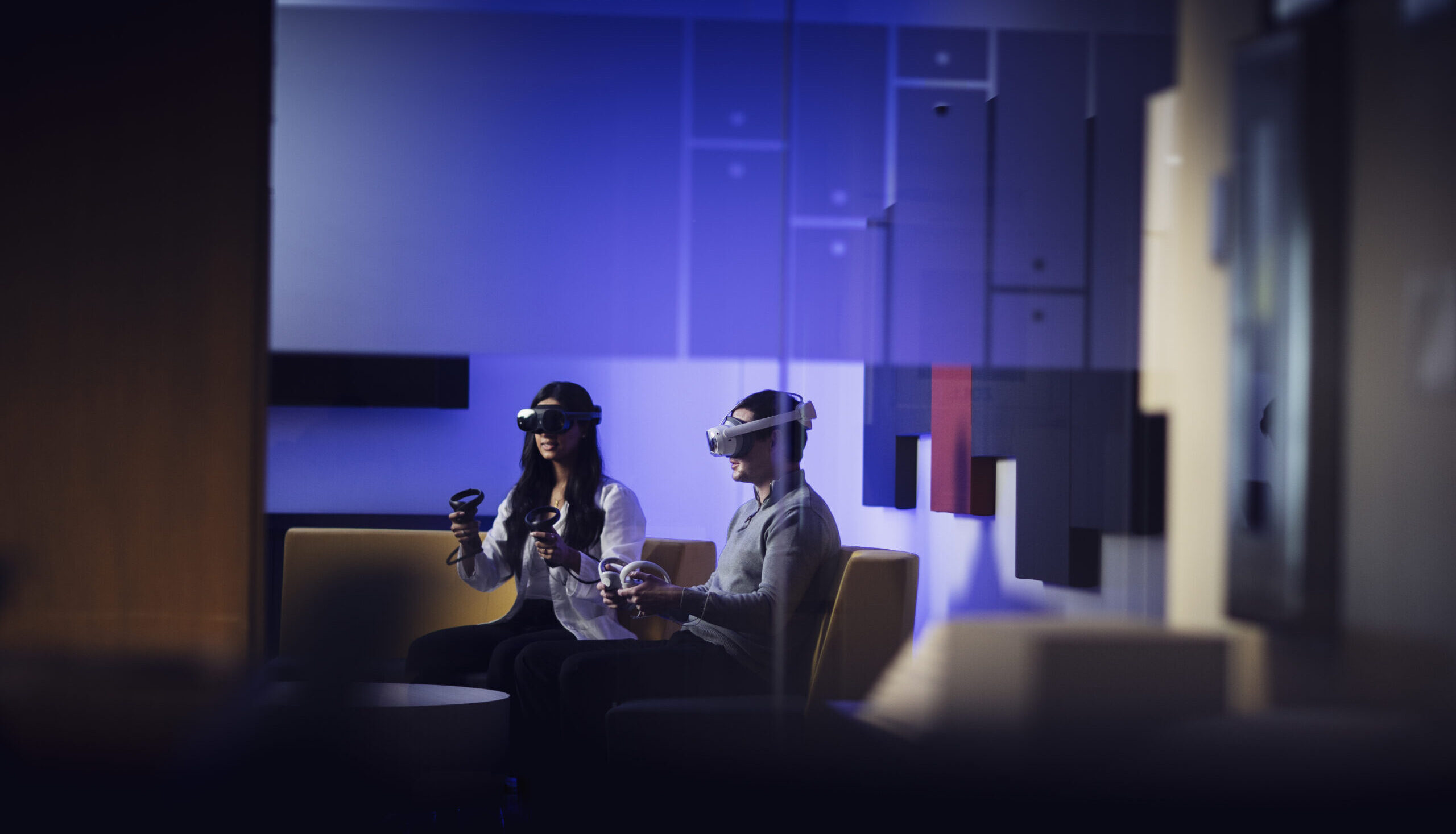 Woman in doctor's coat and man sitting on a couch together, both wearing virtual reality headsets.